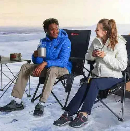 Two people sitting on heated camping chairs outdoors in snowy conditions, enjoying hot drinks during cold-weather camping.