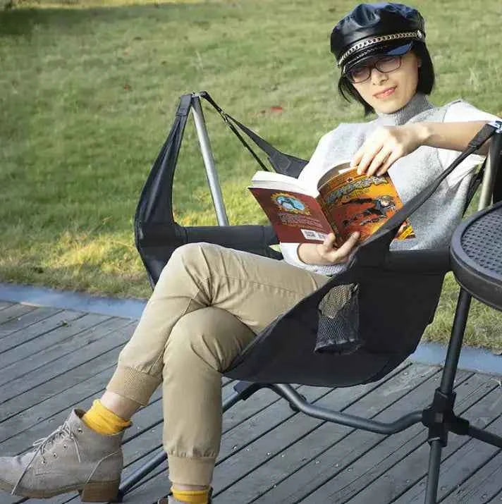 Person relaxing outdoors in a portable hammock chair, seated comfortably while reading a book on a wooden deck.