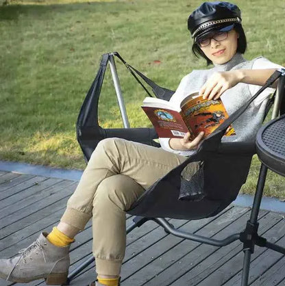 Person relaxing outdoors in a portable hammock chair, seated comfortably while reading a book on a wooden deck.